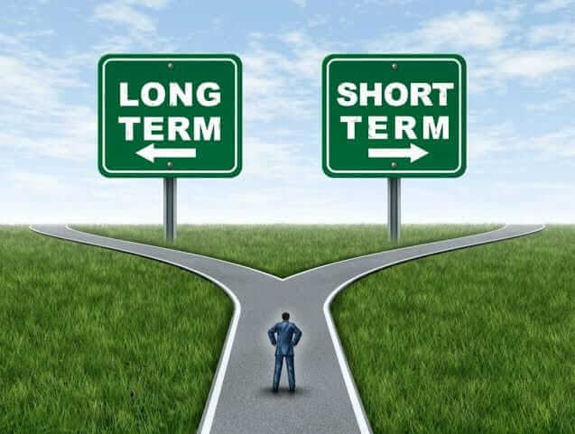 A person standing at a fork in a road, facing two green road signs. The sign to the left points down a path labeled 'LONG TERM', and the sign to the right points down a path labeled 'SHORT TERM