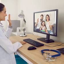 A female doctor consults with a family (a man, woman, and child) via video call on a computer monitor, with a stethoscope resting on the desk in the foreground