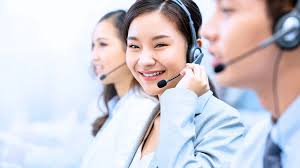 A smiling beautiful Asian woman working in a call center office as a customer service operator, wearing a headset with a microphone, with other call center agents blurred in the background.