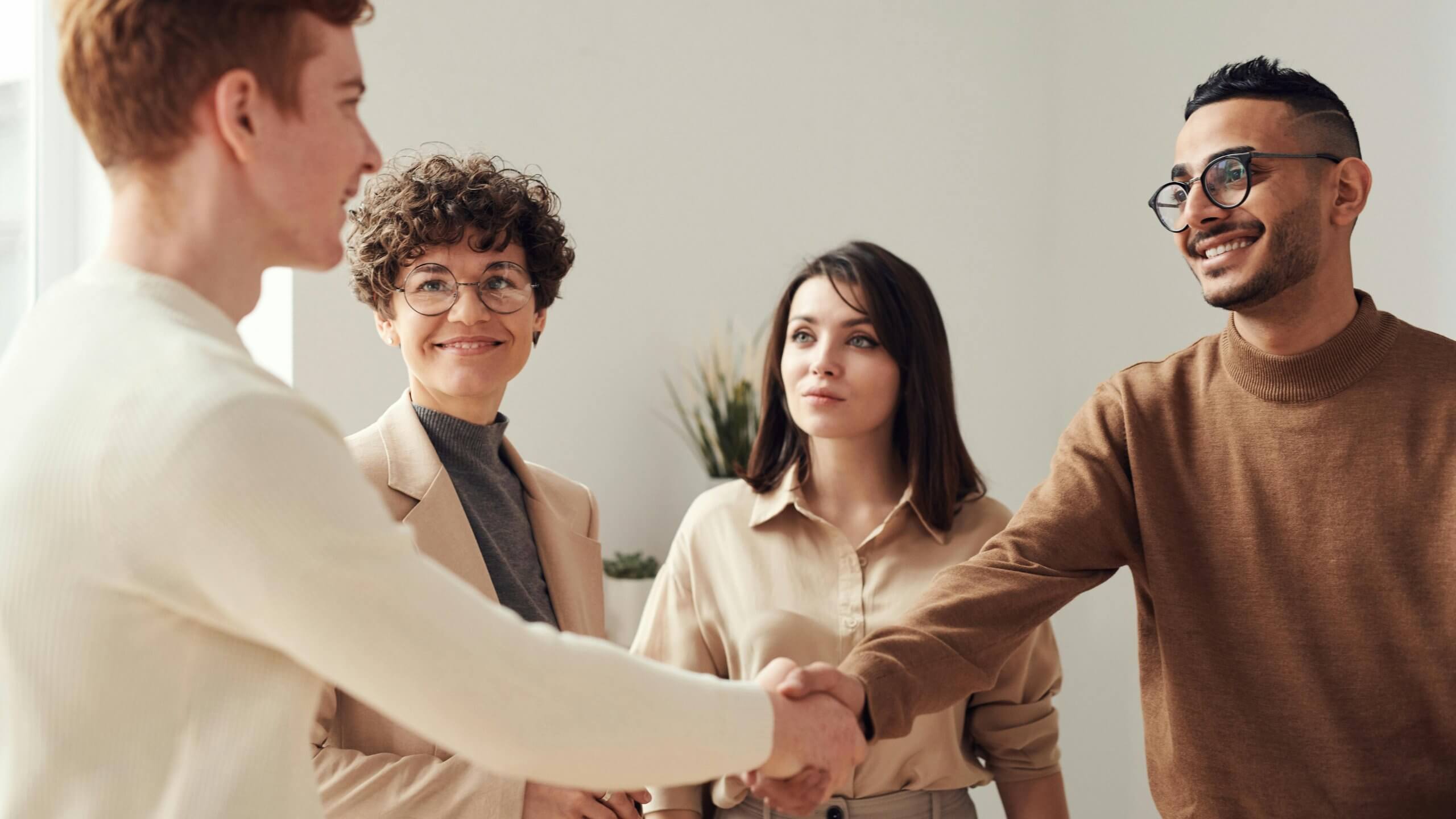 A group of four people, two men and two women, in an office setting. The man on the left, with reddish hair, is shaking hands with the man on the right, who has dark hair and glasses. Two women, one with curly hair and glasses and the other with straight dark hair, are standing behind them, smiling and observing the handshake.