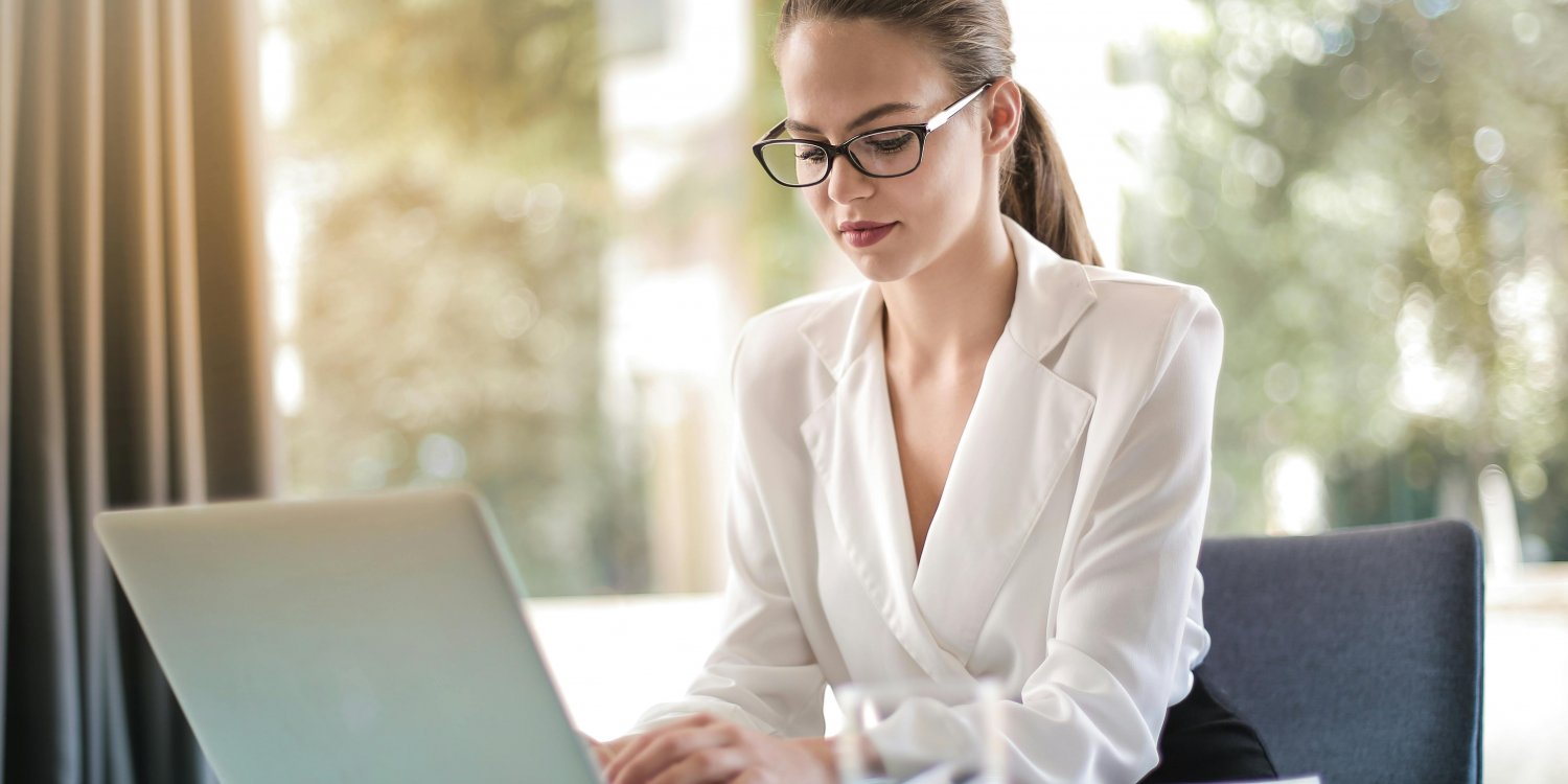 A young woman with glasses and a white blazer works on a laptop at a glass desk in a bright office environment.