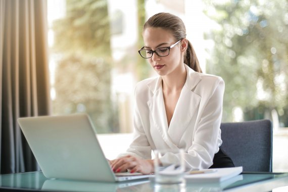 A young woman with glasses and a white blazer works on a laptop at a glass desk in a bright office environment.