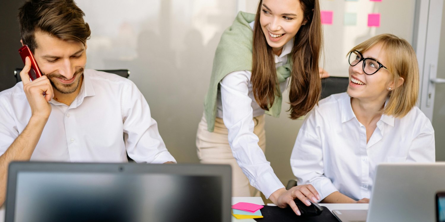Three smiling colleagues working together in a modern office, with one man on the phone, one woman looking at a laptop, and another woman leaning over to interact with the woman at the laptop