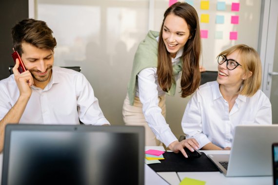 Three smiling colleagues working together in a modern office, with one man on the phone, one woman looking at a laptop, and another woman leaning over to interact with the woman at the laptop