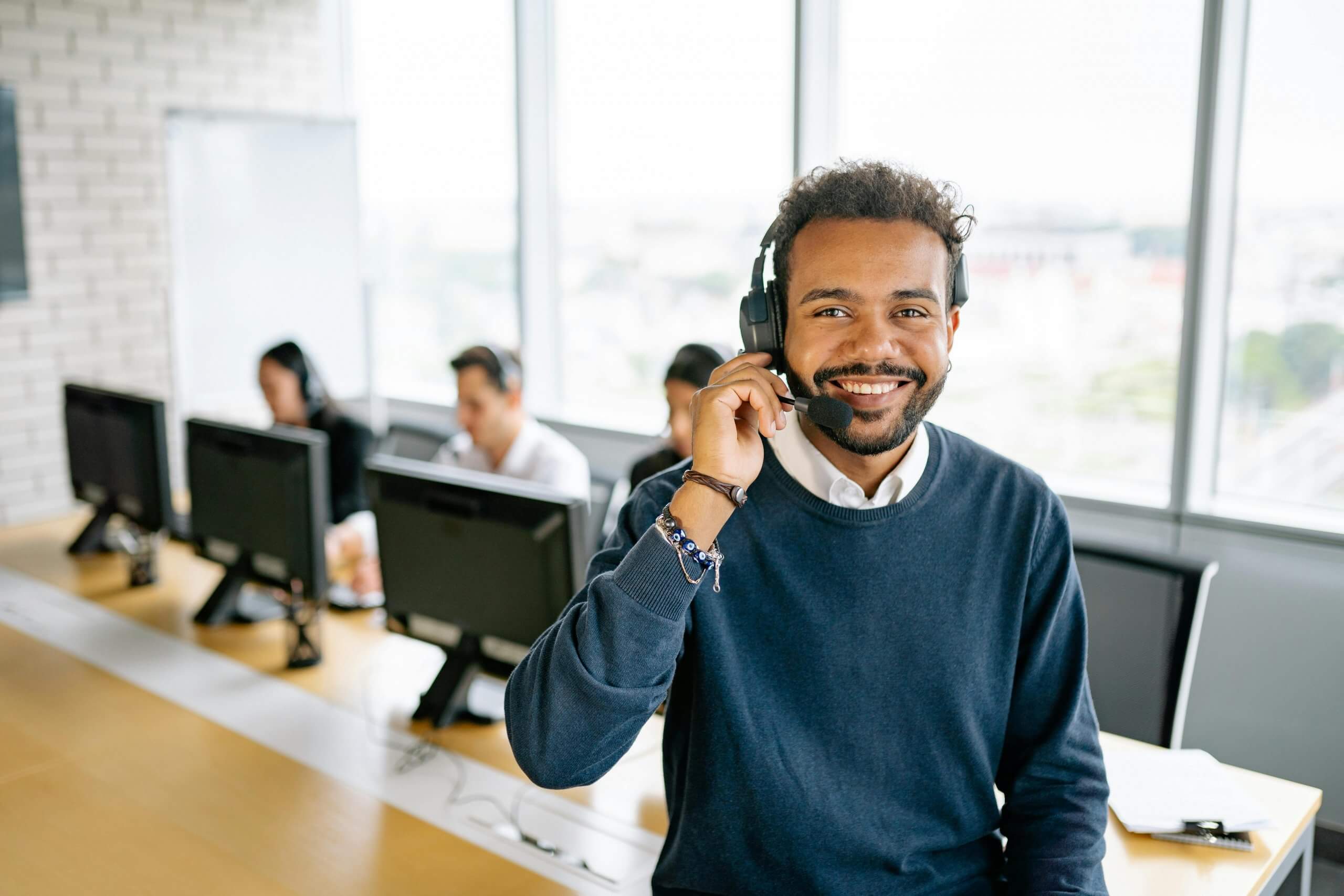A smiling male customer service representative wearing a headset with a microphone, looking directly at the camera in a call center environment