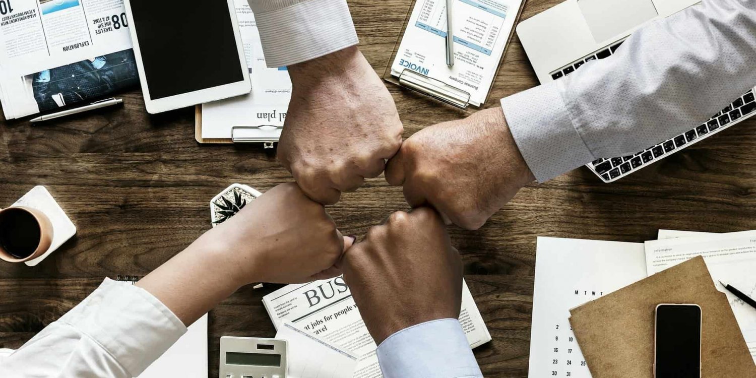 Four diverse people's fists meeting in a huddle over a wooden desk with various business items like a laptop, papers, and a calculator, symbolizing teamwork or collaboration.
