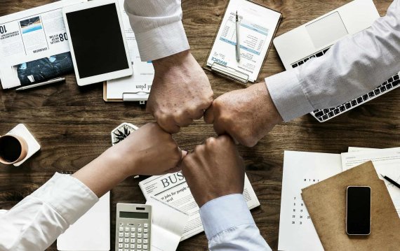 Four diverse people's fists meeting in a huddle over a wooden desk with various business items like a laptop, papers, and a calculator, symbolizing teamwork or collaboration.