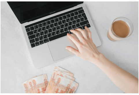A person's hands typing on a silver laptop keyboard with money on the table in the foreground.
