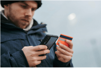 A man in a dark winter coat and hat holds a smartphone in one hand and an orange and grey credit card in the other, appearing to be engaged in an online transaction or checking account details