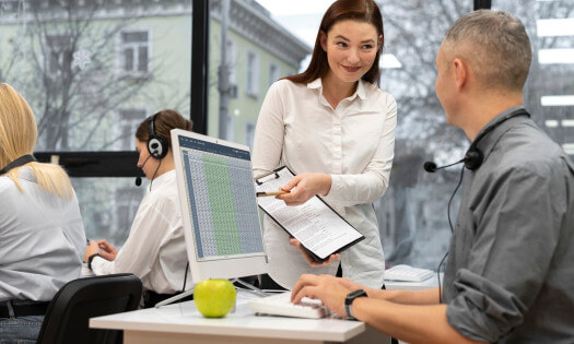 A female call center supervisor assisting a male agent at a desk with a computer and a green apple, while other agents work in the background